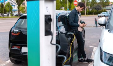 Man charging his electric car at charge station and using smartphone
